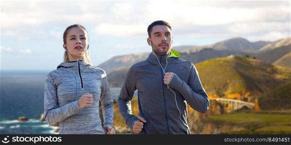 fitness, sport and technology concept - happy couple running and listening to music in earphones over bixby creek bridge on big sur coast of california background. couple with earphones running over big sur coast. couple with earphones running over big sur coast