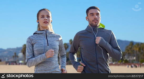 fitness, sport and technology concept - happy couple running and listening to music in earphones over venice beach background in california. couple with earphones running over venice beach. couple with earphones running over venice beach