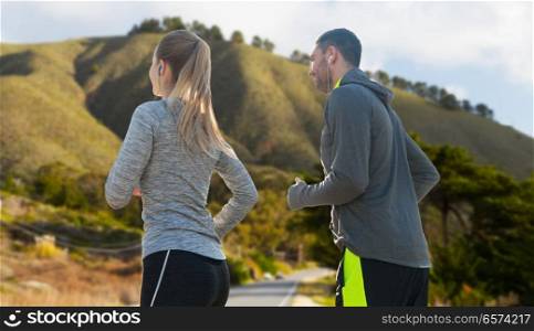 fitness, sport and technology concept - happy couple running and listening to music in earphones over big sur hills background in california. couple with earphones running over big sur