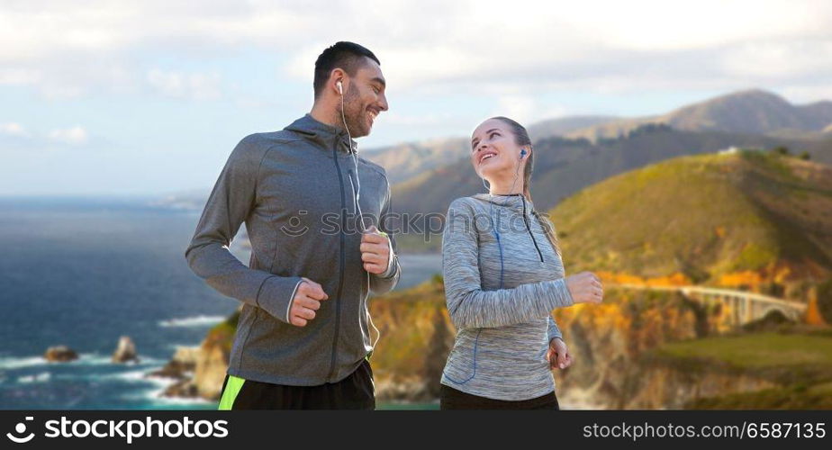 fitness, sport and technology concept - happy couple running and listening to music in earphones over bixby creek bridge on big sur coast of california background. couple with earphones running over big sur coast