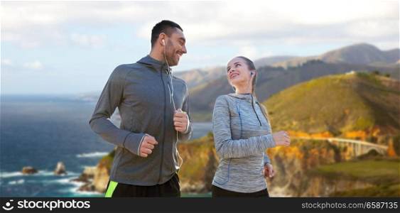 fitness, sport and technology concept - happy couple running and listening to music in earphones over bixby creek bridge on big sur coast of california background. couple with earphones running over big sur coast