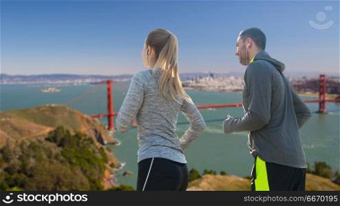 fitness, sport and technology concept - happy couple running and listening to music in earphones over golden gate bridge in san francisco bay background. couple with earphones running over san francisco. couple with earphones running over san francisco