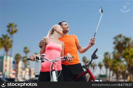 fitness, sport and technology concept - happy couple with bicycle taking picture by smartphone on selfie stick over venice beach background in california. couple with bicycle and smartphone selfie stick. couple with bicycle and smartphone selfie stick
