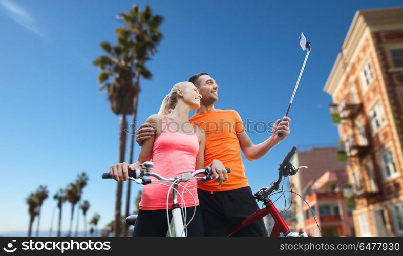 fitness, sport and technology concept - happy couple with bicycle taking picture by smartphone on selfie stick over venice beach background in california. couple with bicycle and smartphone selfie stick. couple with bicycle and smartphone selfie stick