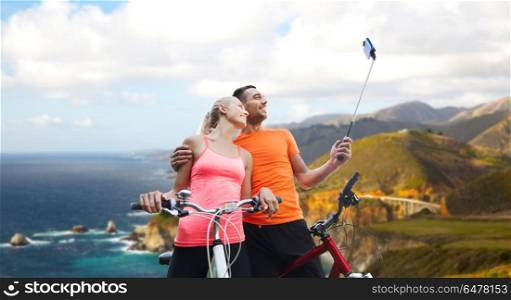 fitness, sport and technology concept - happy couple with bicycle taking picture by smartphone on selfie stick over big sur hills and pacific ocean background in california. couple with bicycle and smartphone selfie stick. couple with bicycle and smartphone selfie stick