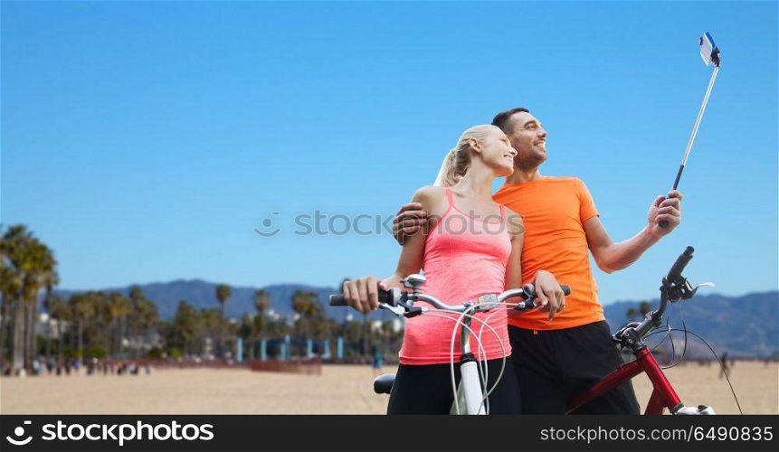 fitness, sport and technology concept - happy couple with bicycle taking picture by smartphone on selfie stick over venice beach background in california. couple with bicycle and smartphone selfie stick. couple with bicycle and smartphone selfie stick