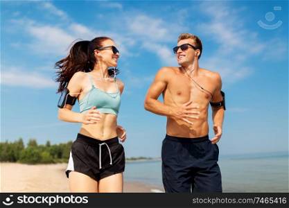 fitness, sport and technology concept - happy couple with earphones and arm bands running along summer beach. couple with phones and arm bands running on beach