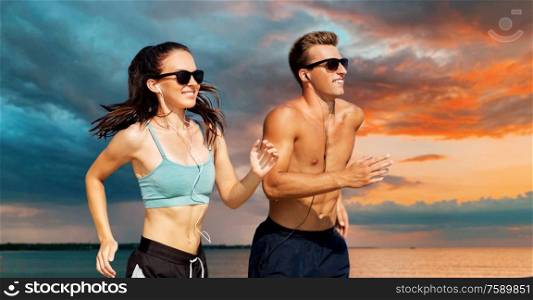 fitness, sport and technology concept - happy couple with earphones running over sea and sunset sky on background. couple with earphones running along on beach
