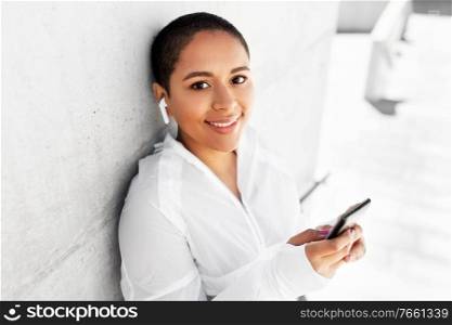 fitness, sport and technology concept - happy smiling young african american woman with wireless earphones listening to music on smartphone outdoors. african american woman with earphones and phone