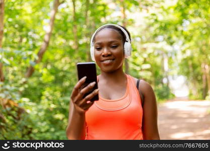 fitness, sport and technology concept - happy smiling young african american woman with headphones listening to music on smartphone outdoors. african american woman with headphones and phone