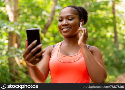 fitness, sport and technology concept - happy smiling young african american woman with earphones listening to music on smartphone outdoors. african american woman with earphones and phone