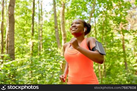 fitness, sport and technology concept - happy smiling young african american woman with earphones and smartphone in arm band running at park. african woman with earphones running at park