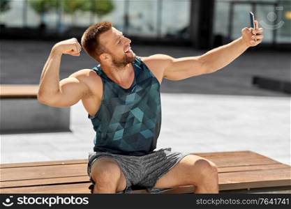 fitness, sport and technology concept - happy smiling young man taking selfie with smartphone sitting on city bench and showing bicep. man taking selfie with phone and showing bicep