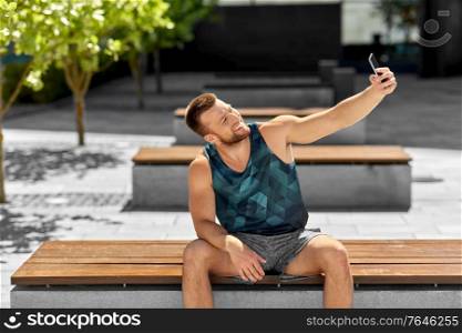 fitness, sport and technology concept - happy smiling young man taking selfie with smartphone sitting on bench outdoors. man taking selfie with smartphone outdoors