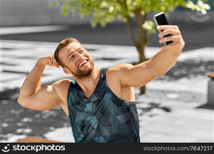 fitness, sport and technology concept - happy smiling young man taking selfie with smartphone sitting on city bench and showing bicep. man taking selfie with phone and showing bicep