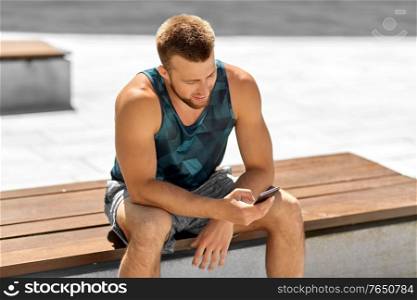 fitness, sport and technology concept - happy smiling young man with smartphone sitting on bench outdoors. man with smartphone sitting on bench outdoors