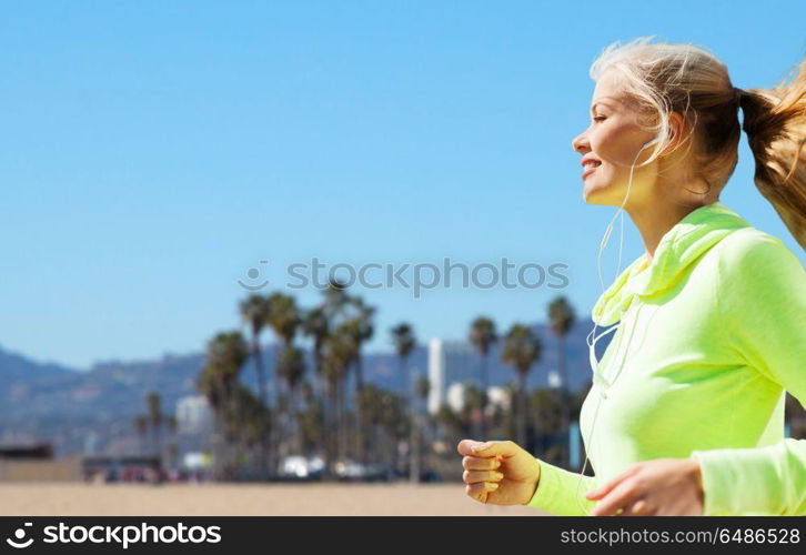fitness, sport and technology concept - happy woman running and listening to music in earphones over venice beach background in california. woman with earphones running over venice beach. woman with earphones running over venice beach