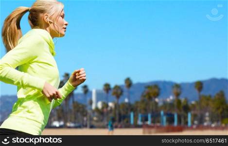 fitness, sport and technology concept - happy woman running and listening to music in earphones over venice beach background in california. woman with earphones running over venice beach. woman with earphones running over venice beach