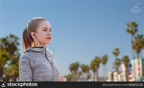 fitness, sport and technology concept - happy woman running and listening to music in earphones over venice beach background in california. woman with earphones running over venice beach