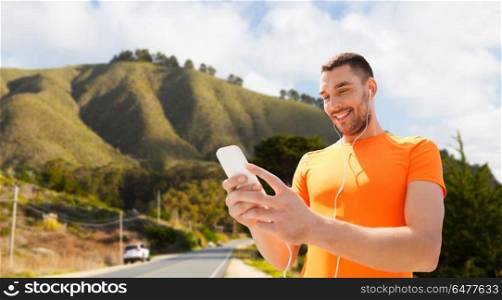 fitness, sport and technology concept - smiling man with smartphone and earphones listening to music over big sur hills and road background in california. man with smartphone and earphones over hills. man with smartphone and earphones over hills