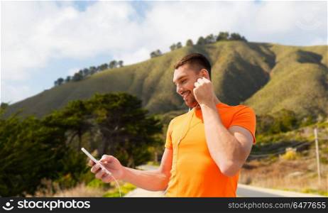 fitness, sport and technology concept - smiling man with smartphone and earphones listening to music over big sur hills and road background in california. man with smartphone and earphones over hills. man with smartphone and earphones over hills