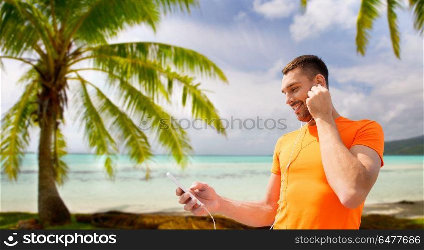 fitness, sport and technology concept - smiling man with smartphone and earphones listening to music over tropical beach background in french polynesia. man with smartphone and earphones over beach. man with smartphone and earphones over beach