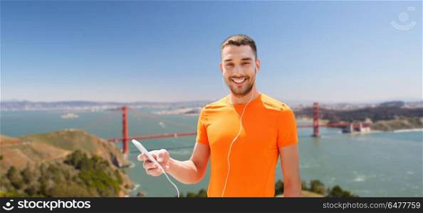 fitness, sport and technology concept - smiling man with smartphone and earphones listening to music over golden gate bridge in san francisco bay background. man with smartphone and earphones over golden gate. man with smartphone and earphones over golden gate