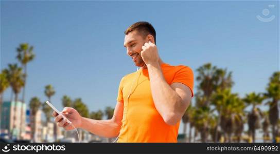 fitness, sport and technology concept - smiling man with smartphone and earphones listening to music over venice beach background in california. man with smartphone and earphones in los angeles. man with smartphone and earphones in los angeles