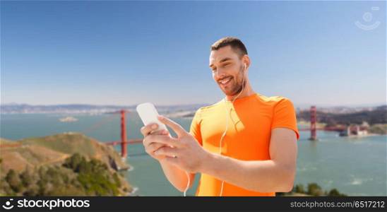 fitness, sport and technology concept - smiling man with smartphone and earphones listening to music over golden gate bridge in san francisco bay background. man with smartphone and earphones over golden gate. man with smartphone and earphones over golden gate