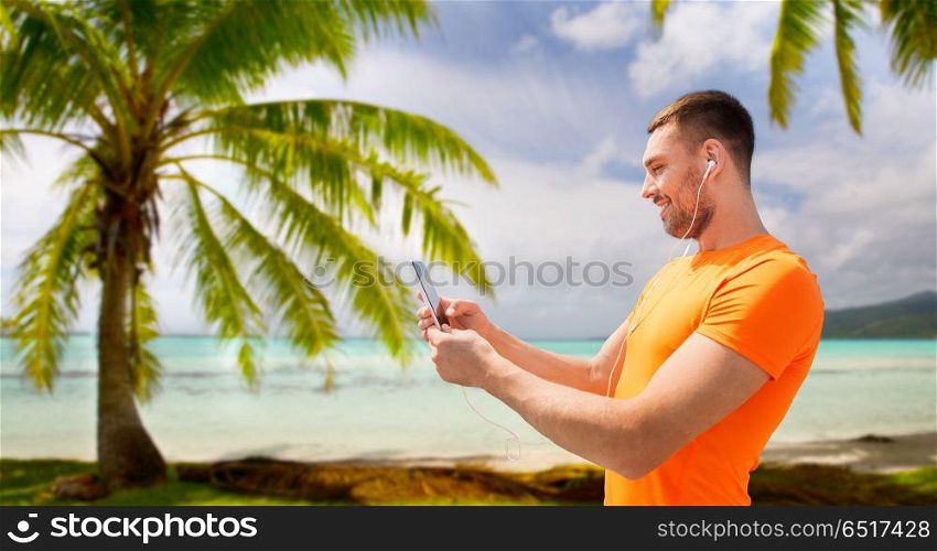 fitness, sport and technology concept - smiling young man with smartphone and earphones listening to music over tropical beach background in french polynesia. smiling young man with smartphone and earphones. smiling young man with smartphone and earphones