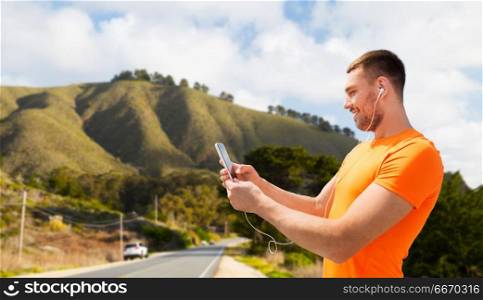 fitness, sport and technology concept - smiling young man with smartphone and earphones listening to music over big sur hills and road background in california. man with smartphone and earphones over big sur. man with smartphone and earphones over big sur