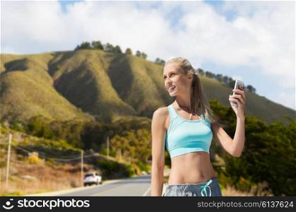 fitness, sport and technology concept - smiling young woman with smartphone exercising over big sur hills and road background in california. woman with smartphone doing sports over hills. woman with smartphone doing sports over hills