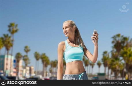 fitness, sport and technology concept - smiling young woman with smartphone exercising over venice beach background in california. woman with smartphone doing sports in los angeles. woman with smartphone doing sports in los angeles