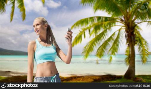 fitness, sport and technology concept - smiling young woman with smartphone over tropical beach background in french polynesia. woman with smartphone doing sports over beach. woman with smartphone doing sports over beach