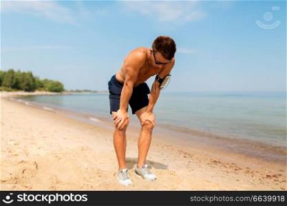 fitness, sport and technology concept - tired male runner with earphones and smartphone in arm band on beach. male runner with earphones and arm band on beach