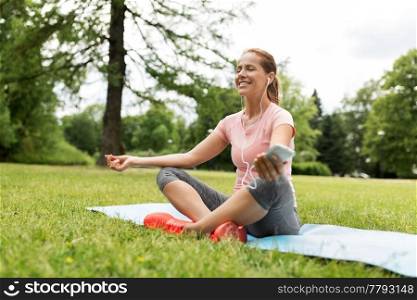 fitness, sport and technology concept - woman with smartphone and earphones meditating on yoga mat at park. woman meditating on yoga mat at park