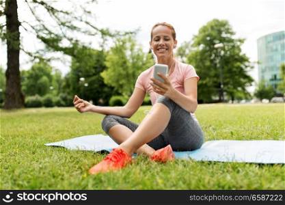 fitness, sport and technology concept - woman with smartphone and earphones resting on exercise mat at park. woman with smartphone resting on mat at park
