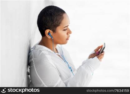 fitness, sport and technology concept - young african american woman with earphones listening to music on smartphone outdoors. african american woman with earphones and phone