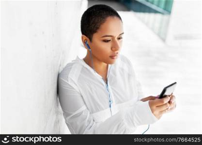 fitness, sport and technology concept - young african american woman with earphones listening to music on smartphone outdoors. african american woman with earphones and phone