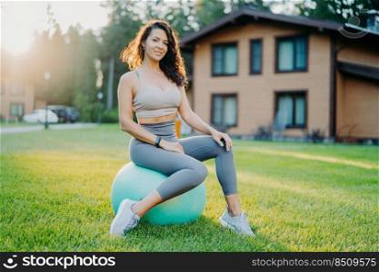 Fitness, sport and training concept. Satisfied sporty woman in good physical shape takes break after exercising sits on fitness ball outdoor enjoys nature and fresh air, does pilates balance exercises