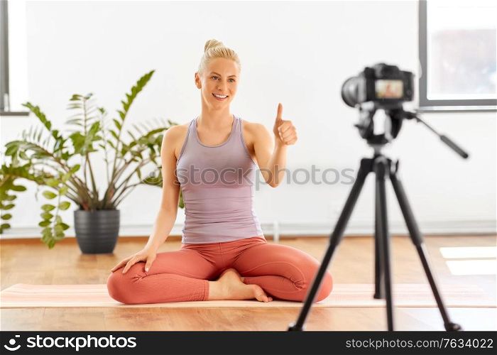 fitness, sport and video blogging concept - happy smiling young woman or blogger with camera on tripod recording online yoga class in gym or studio and showing thumbs up. woman with camera streaming for yoga blog at home