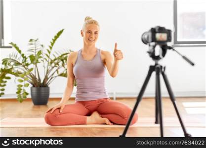 fitness, sport and video blogging concept - happy smiling young woman or blogger with camera on tripod recording online yoga class in gym or studio and showing thumbs up. woman with camera streaming for yoga blog at home