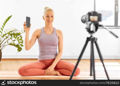 fitness, sport and video blogging concept - happy smiling young woman or blogger with smartphone and camera on tripod recording online yoga class at home or studio. woman with phone streaming for yoga blog at home