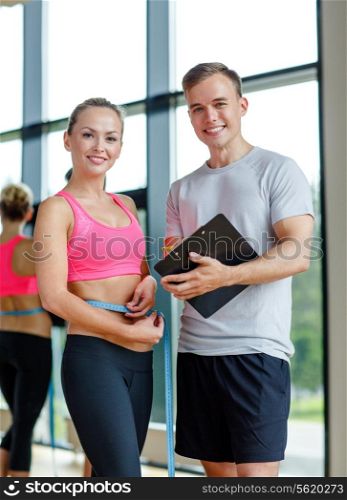 fitness, sport, exercising and diet concept - smiling young woman with measuring tape and personal trainer in gym