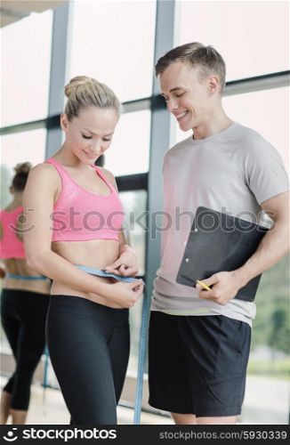 fitness, sport, exercising and diet concept - smiling young woman with measuring tape and personal trainer in gym