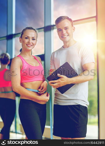 fitness, sport, exercising and diet concept - smiling young woman with measuring tape and personal trainer in gym. smiling young woman with personal trainer in gym