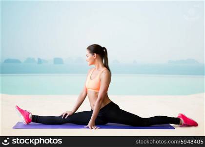 fitness, sport, exercising, stretching and people concept - smiling woman doing splits on mat over sea and pool at hotel resort background