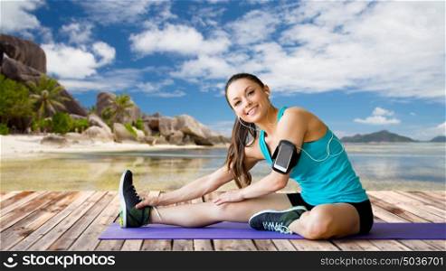 fitness, sport, exercising, technology and people concept - smiling woman wearing armband with smartphone and earphones stretching leg on mat over exotic tropical beach background. woman with smartphone stretching leg on mat