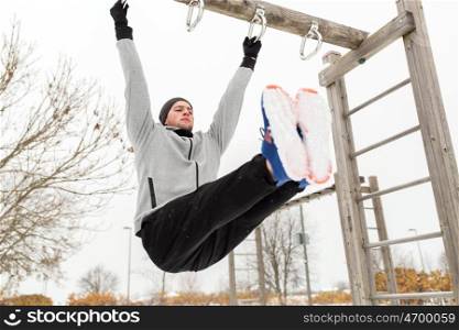 fitness, sport, exercising, training and people concept - young man doing leg pull ups on horizontal bar and flexing abdominal muscles outdoors in winter