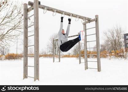 fitness, sport, exercising, training and people concept - young man doing leg pull ups on horizontal bar and flexing abdominal muscles outdoors in winter